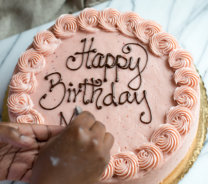 Hand-decorating a custom pink birthday cake with 'Happy Birthday' chocolate lettering at Mad Hatter Cafe & Bakeshop in Durham