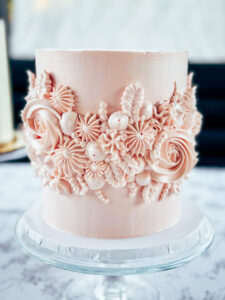 Soft pink buttercream wedding cake with intricate floral details on a glass stand.