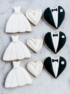 An assortment of wedding dress and tuxedo themed cookies tastefully displayed on a marble countertop.